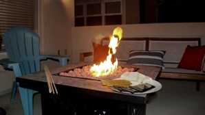 A fire pit with wood logs, surrounded by a table with a plate of food and utensils. There are chairs and a sofa in the background. - Multi Family Home 2 min walk to beach, Sleeps 13 (Oceano)