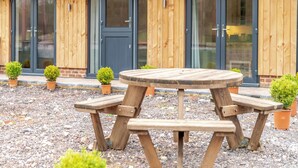 Ein Holzgebäude mit großen Glasfenstern und -türen, eine Schotterterrasse mit einem Holztisch und Bänken sowie kleine Kübelpflanzen.
