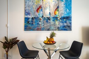 Un comedor con una mesa de vidrio, dos sillas negras y un frutero. Un cuadro abstracto de gran tamaño adorna la pared.