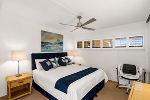 Un dormitorio ordenado con una cama, dos mesitas de noche con lámparas, un ventilador de techo, un escritorio con silla y una ventana con persianas.