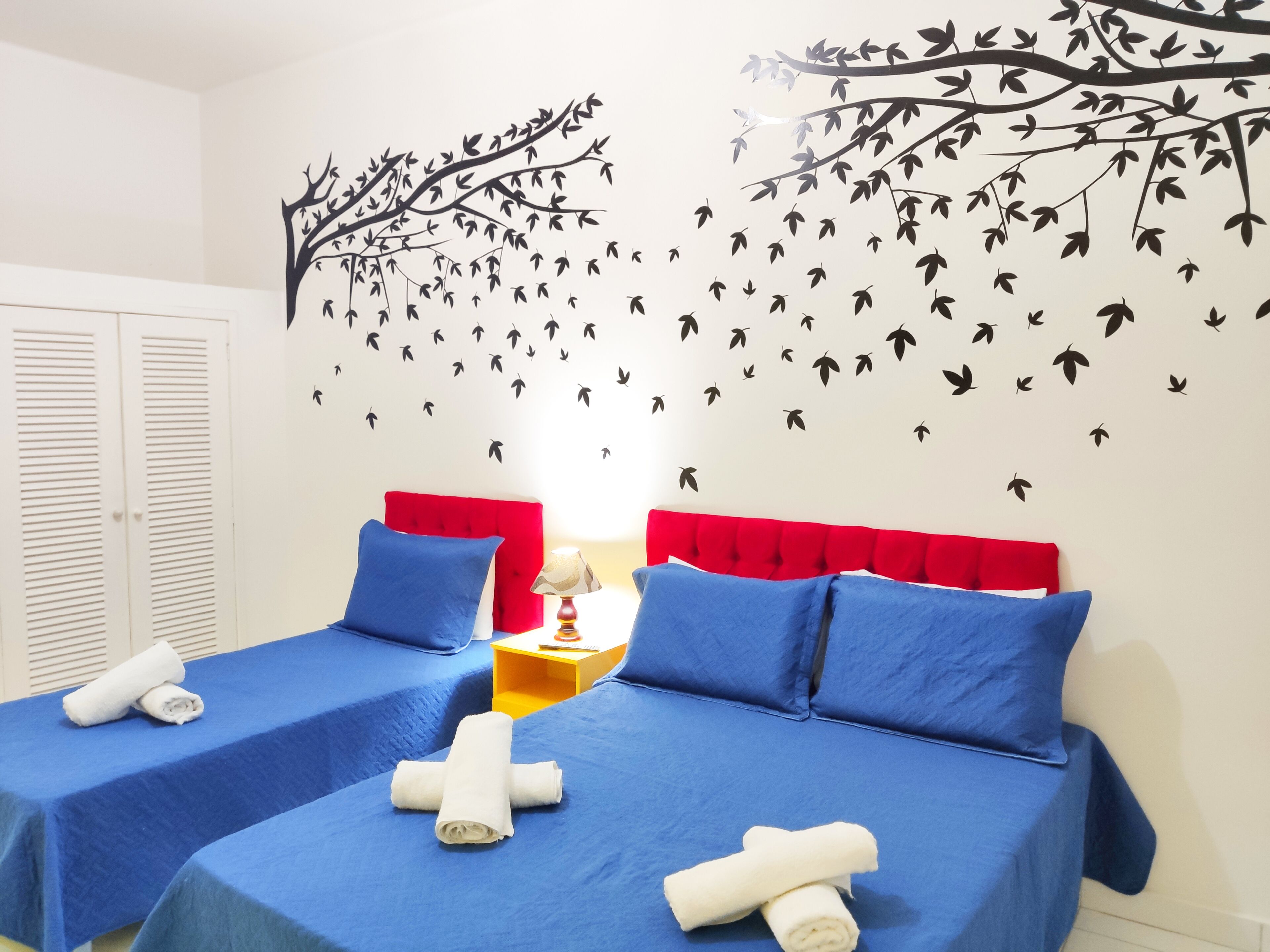 Une chambre d’hôtel avec deux lits, chacun doté de draps bleus et d’une tête de lit rouge. Des serviettes sont posées sur les lits, et un motif décoratif représentant des oiseaux et des branches orne le mur.