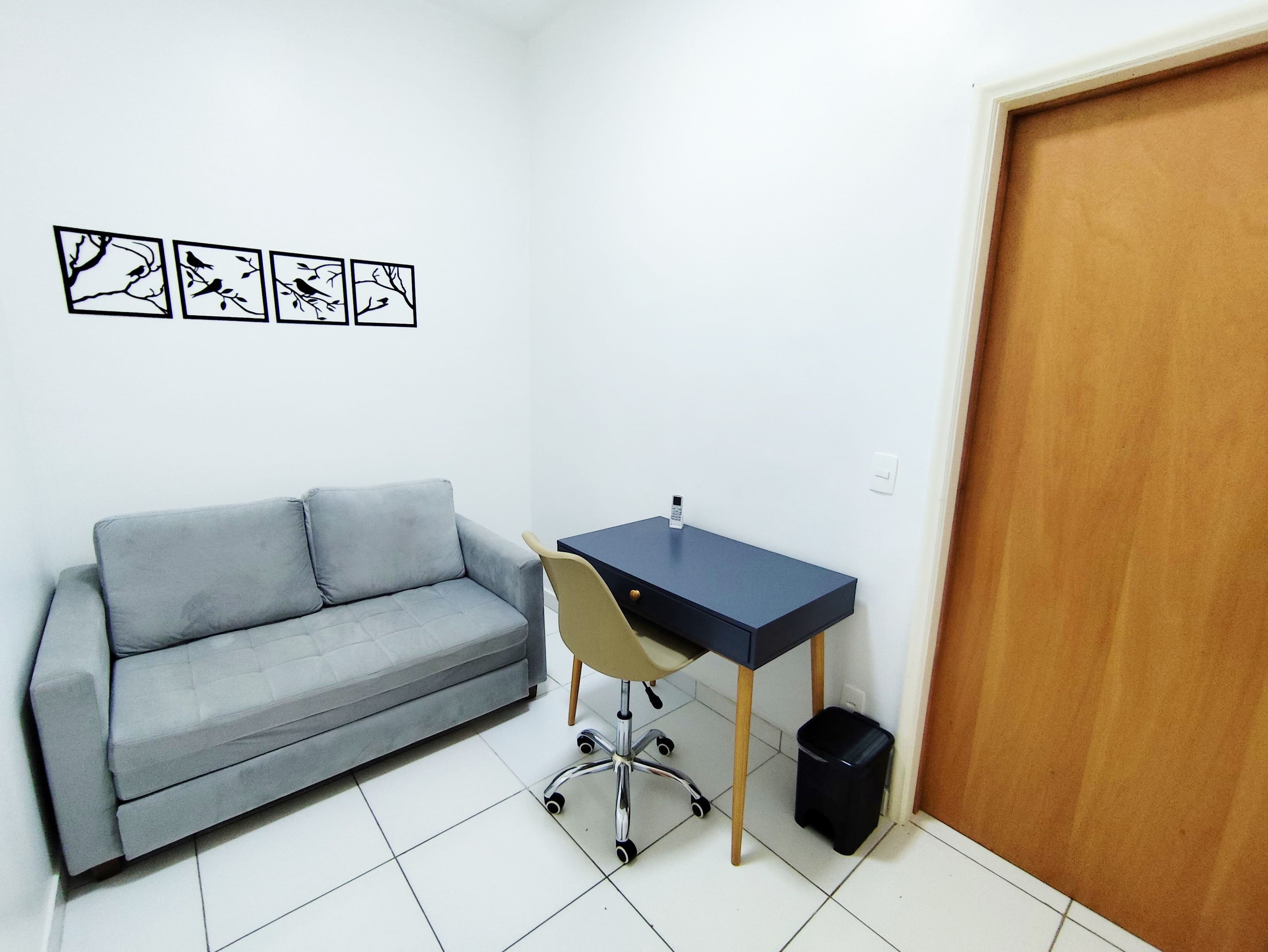 A small, clean room with a grey sofa, a desk with a chair, and a black trash bin. There are four framed pictures on the wall.