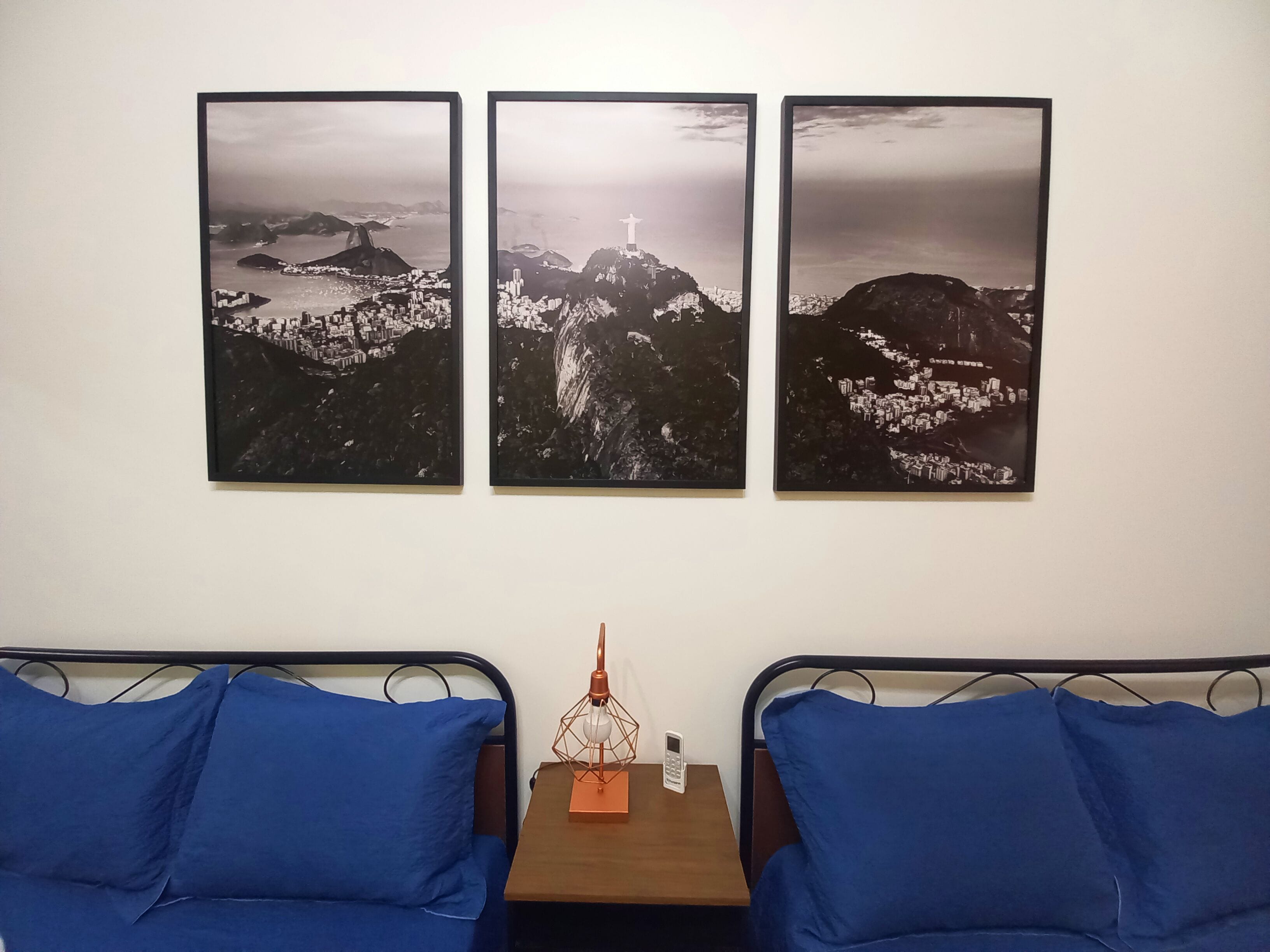Una habitación de hotel con una cama, dos almohadas azules, una mesita de noche con una lámpara y tres cuadros con vistas a un paisaje urbano.