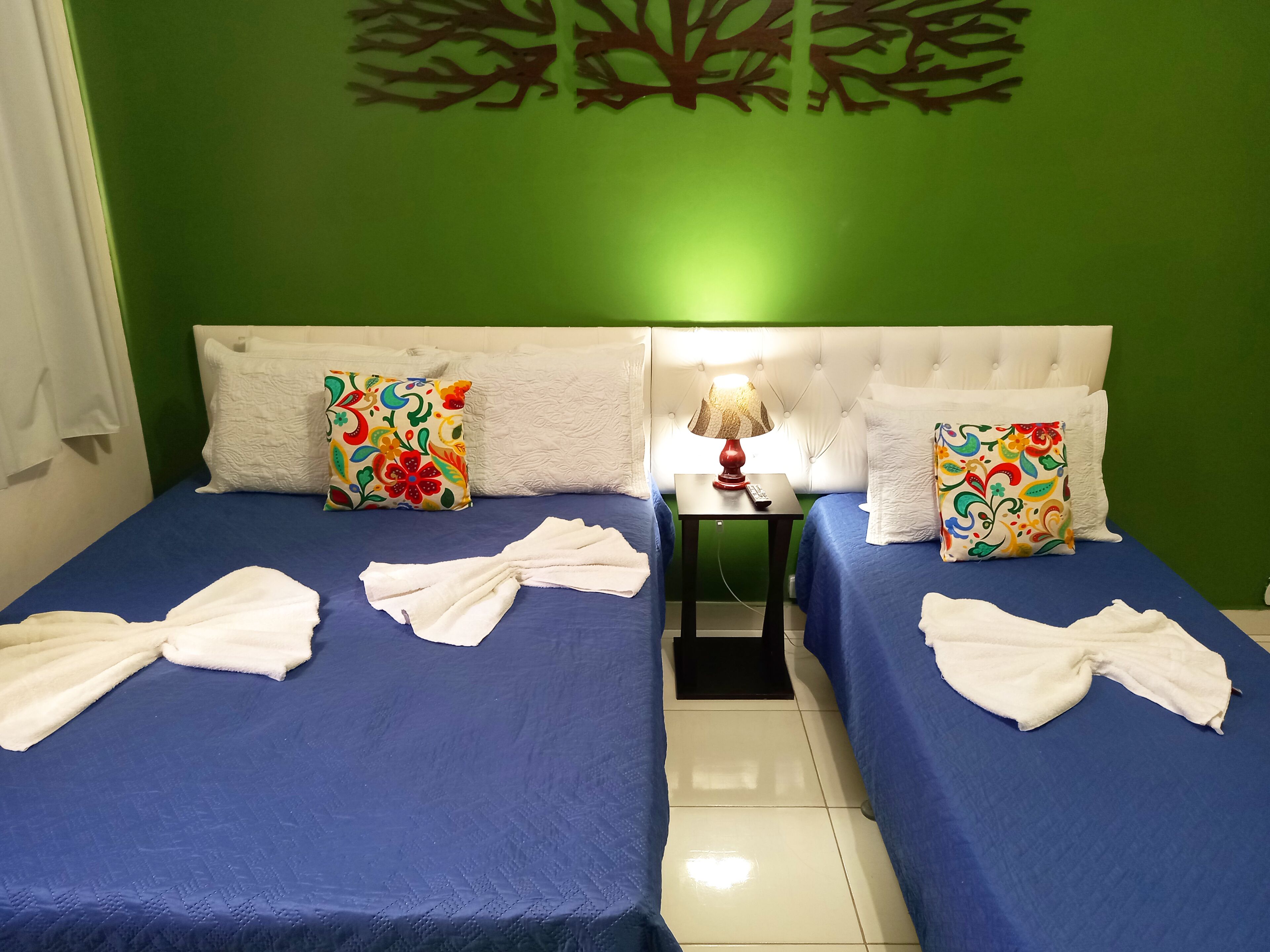 A hotel room with two beds, each with a blue bedspread and white towels. There is a nightstand with a lamp and a remote control between the beds. The walls are green, and there is a decorative tree design above the beds.