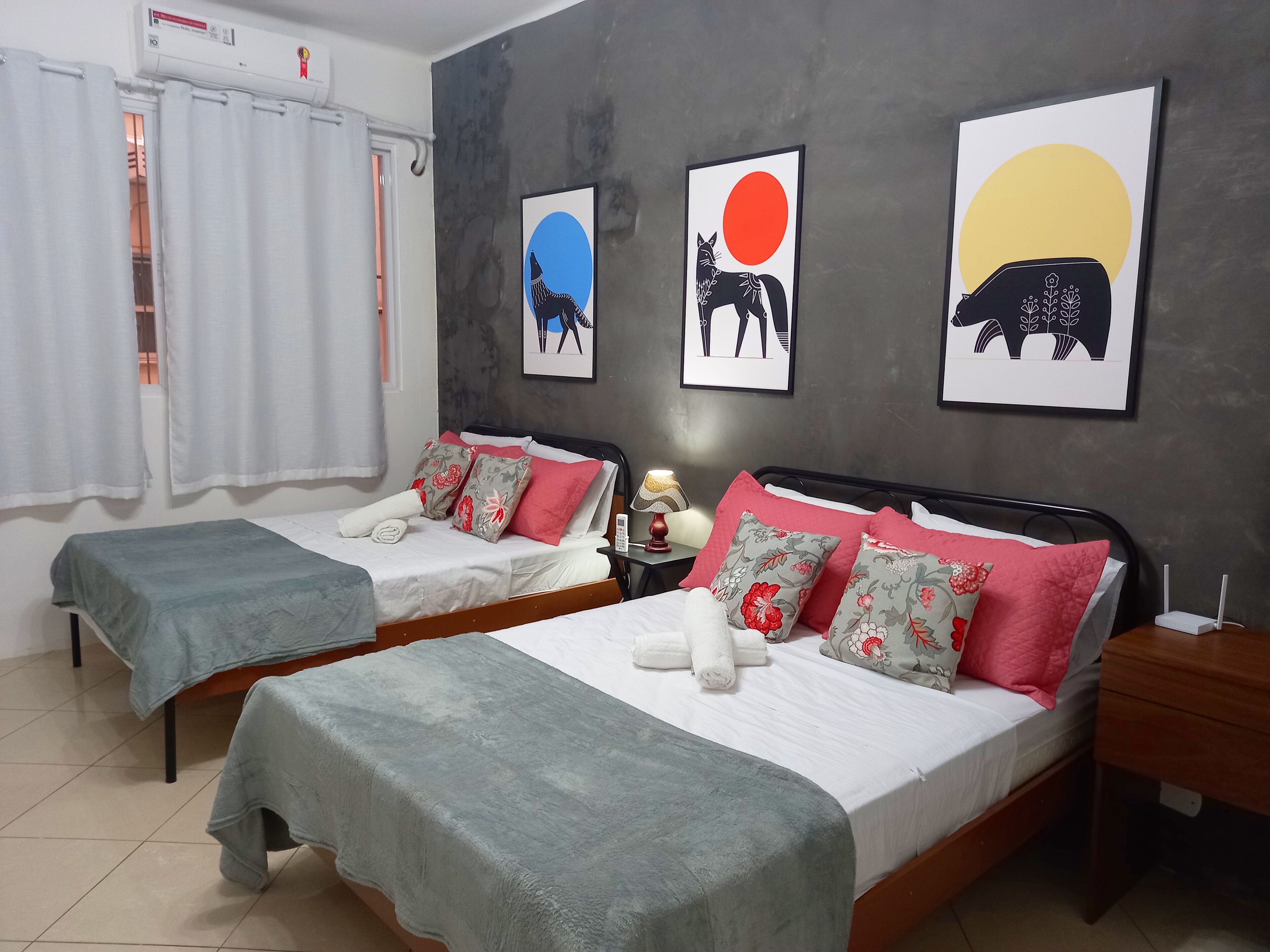 A hotel room with two beds, each with a grey blanket and pink and floral pillows. There are framed artworks on the wall depicting animals and a sun.