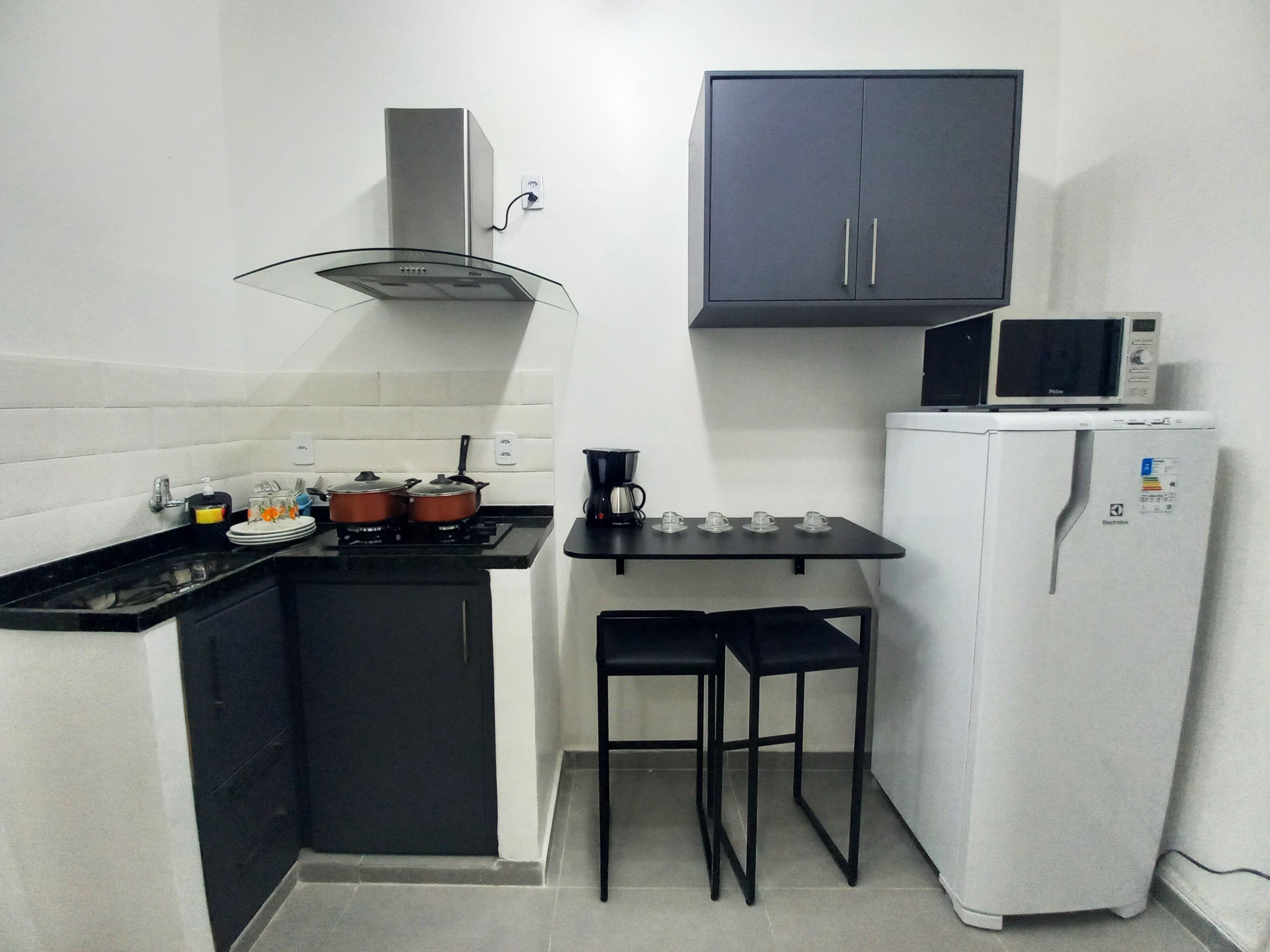 A compact kitchen with a gas stove, two black stools, a white refrigerator, and a microwave on top.