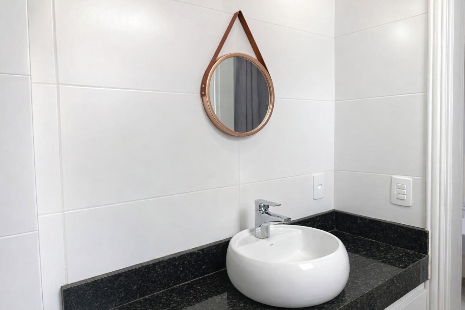 A bathroom with white tiles, a round mirror with a brown frame, a white sink, and a black countertop.