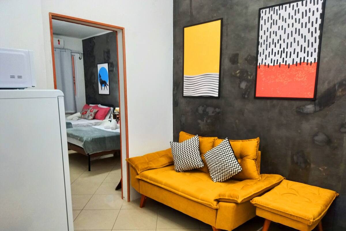 A room with a yellow sofa, a matching ottoman, and a bed with a grey and white duvet. There are two framed artworks on the wall, one with a yellow background and the other with a black and white pattern.