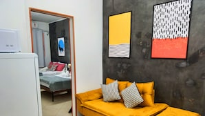 Una habitación con un sofá amarillo, un banquillo a juego y una cama con edredón gris y blanco. Hay dos cuadros en la pared, uno con fondo amarillo y el otro con diseño en blanco y negro.