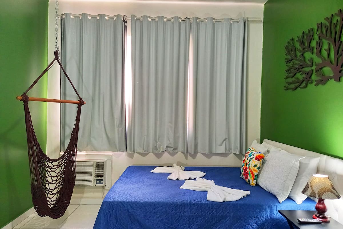 A hotel room with a bed, a hanging chair, a window with curtains, and a wall with a tree decoration.
