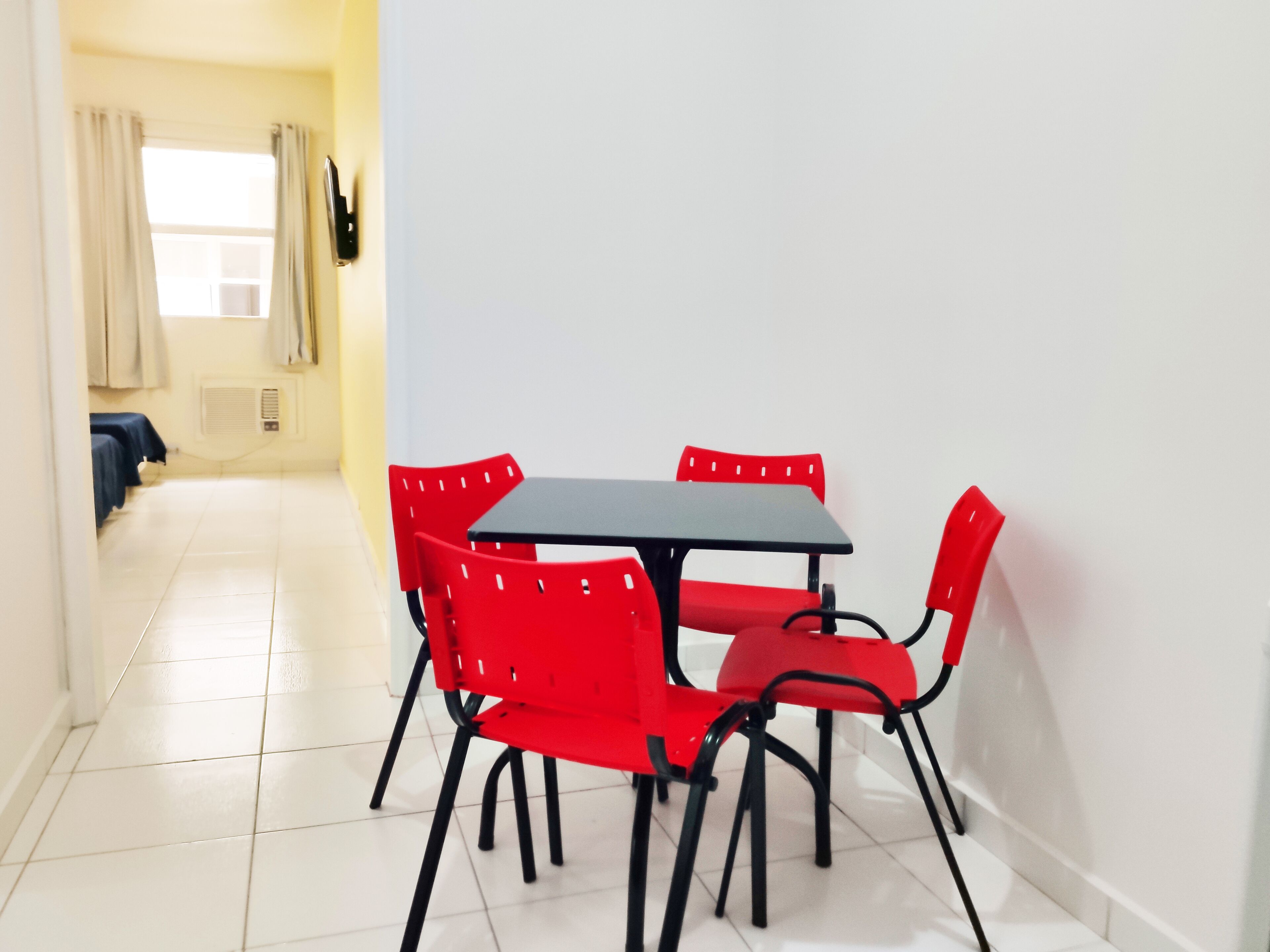 Ein Zimmer mit Fliesenboden, einem schwarzen Tisch und roten Stühlen. Im Hintergrund ist ein Fenster mit Vorhängen und eine Tür zu sehen.