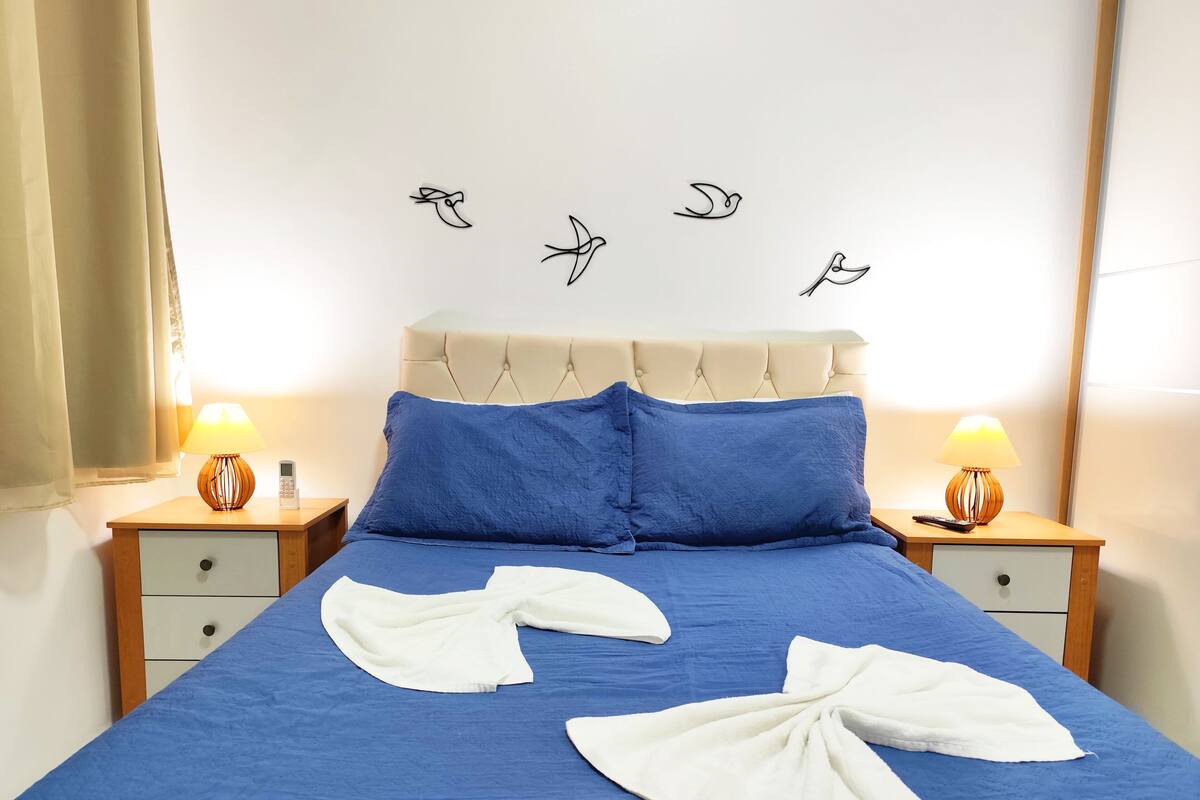 A neatly made bed with blue bedding and white towels, a beige headboard, and two bedside tables with lamps.