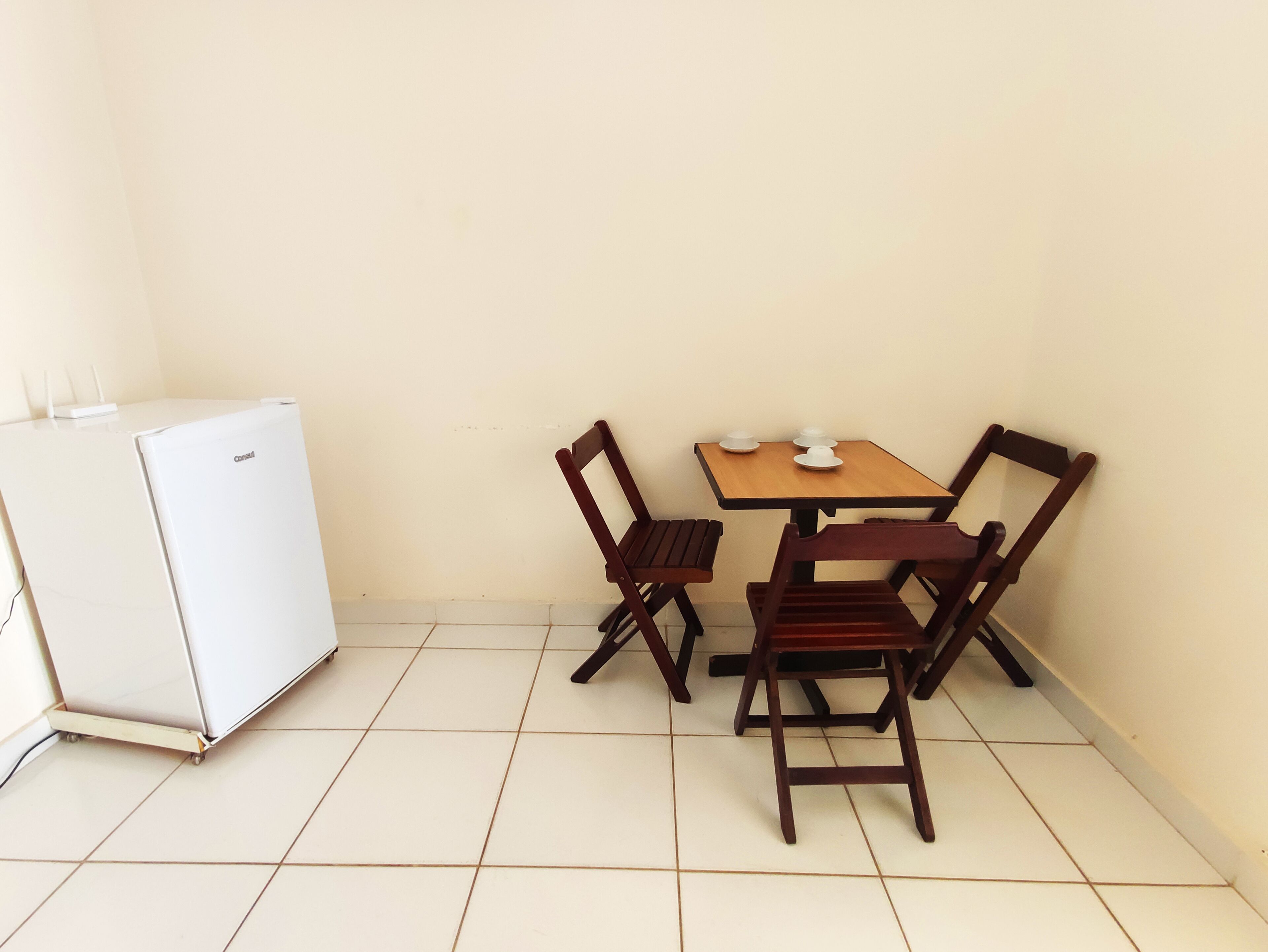 Une petite pièce avec une table en bois et quatre chaises, un réfrigérateur blanc et un sol carrelé.