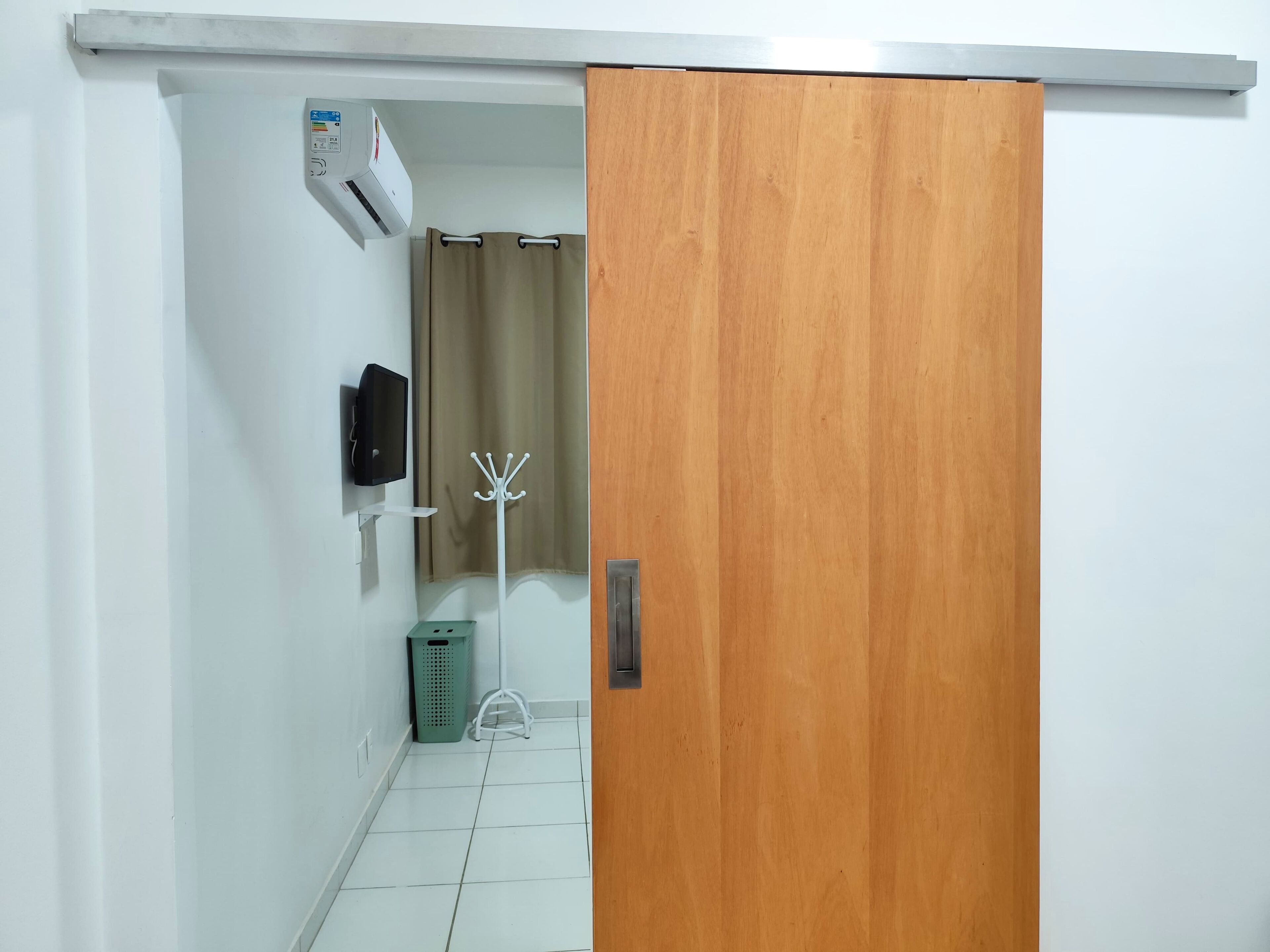 A hallway with a wooden door, a wall-mounted air conditioner, a television, a coat rack, a trash bin, and a small white stool.