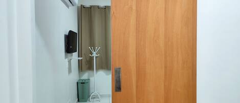 A hallway with a wooden door, a wall-mounted air conditioner, a television, a coat rack, a trash bin, and a small white stool.