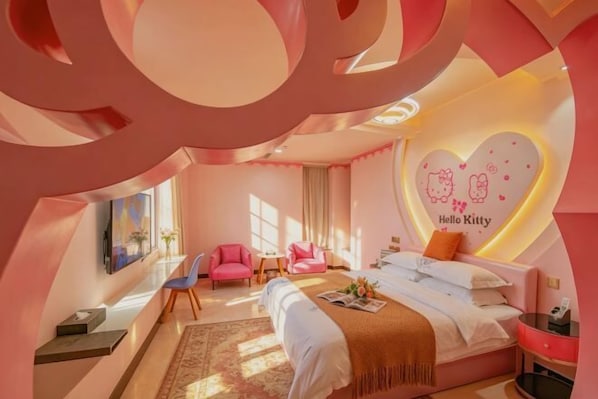 Une chambre d’hôtel décorée dans des tons roses, comprenant un lit, deux chaises, un bureau et un mur orné d’un motif Hello Kitty.