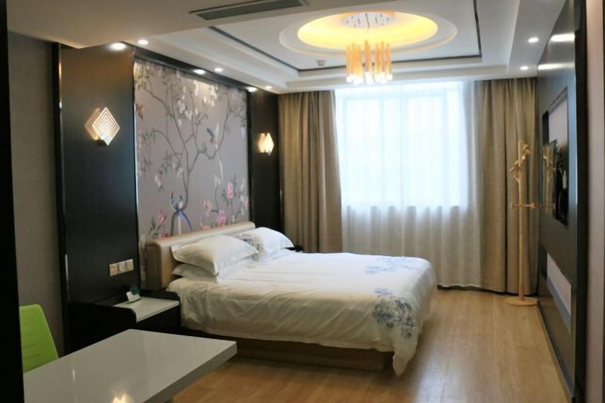 A modern hotel room with a large bed, a bedside table, a desk, and a large window with curtains.