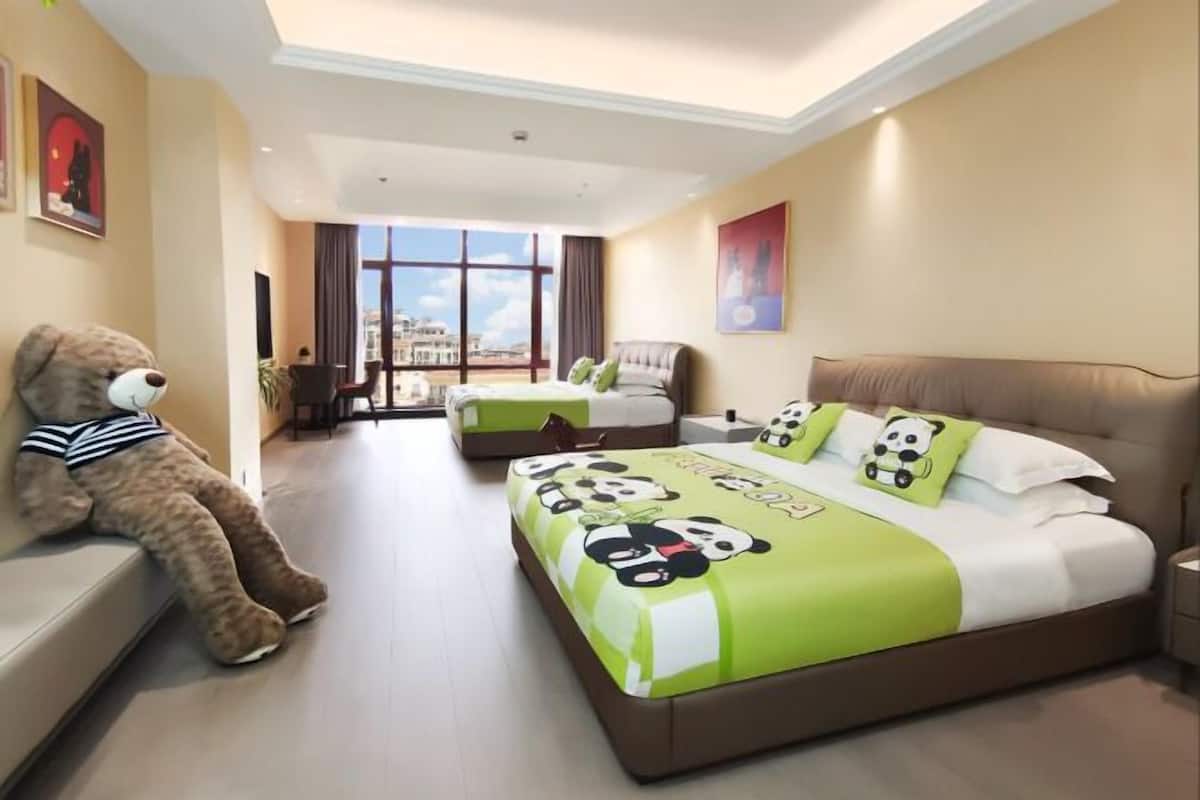 Habitación de hotel con dos camas, un ventanal con vista a la ciudad, un osito de peluche en un banco y obras de arte en las paredes.