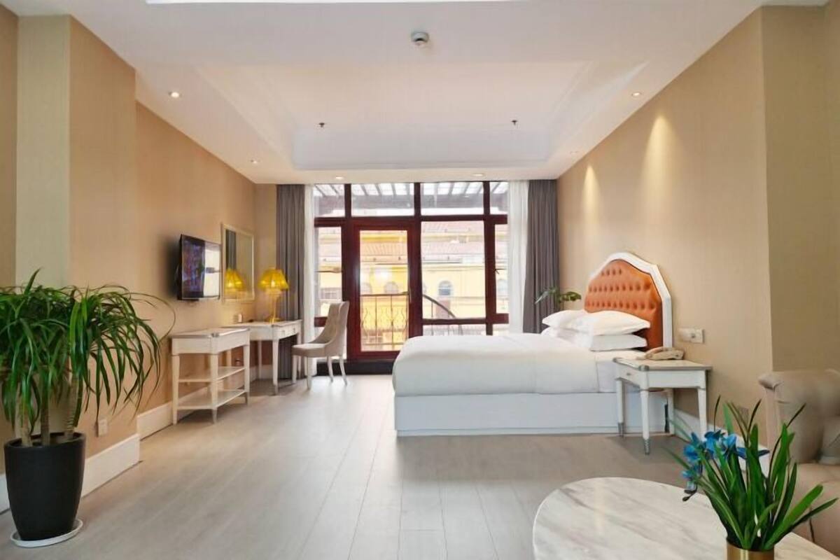 Habitación de hotel con cama, escritorio, silla, plantas en macetas y un ventanal amplio.