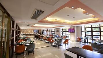 A spacious restaurant with multiple dining tables and chairs, a bar area, and large windows providing natural light.