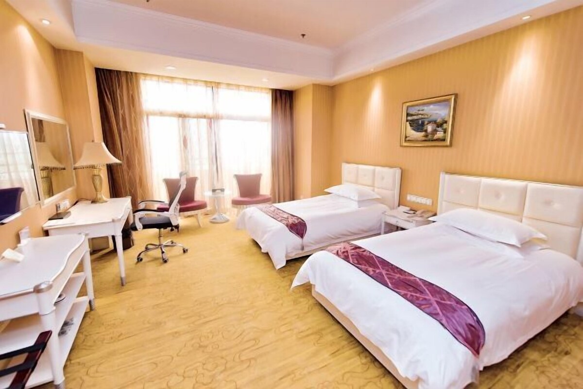 Ein Hotelzimmer mit zwei Betten, einem Schreibtisch, einem Stuhl und einem großen Fenster mit Vorhängen.