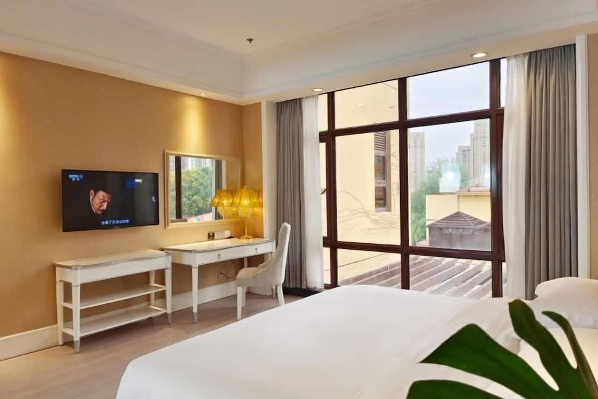 Habitación de hotel con cama, escritorio, televisión, un ventanal con cortinas y vistas a edificios.