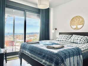 Ein Schlafzimmer mit großem Fenster, einem Bett mit blau-weißer Bettdecke, einer an der Wand befestigten Baumdekoration und Blick auf eine Stadtlandschaft.