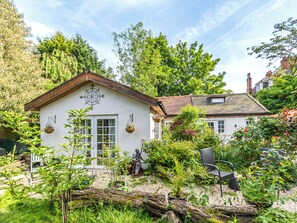 Une petite maison blanche avec une boussole sur le mur avant, entourée d’une végétation luxuriante et d’un jardin où poussent différentes plantes, ainsi qu’une statue.