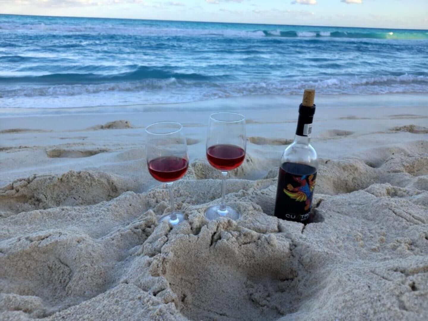 A bottle of wine and two glasses on a sandy beach with the ocean in the background.