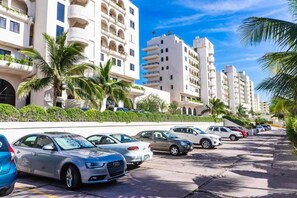 A row of white multi-story buildings with balconies, parked cars, and palm trees.