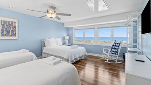 A bedroom with two beds, a ceiling fan, a window with a view of the beach, and a white rocking chair.