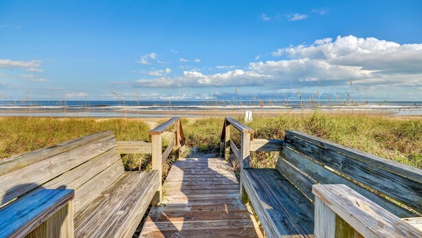 A wooden boardwalk leading to a beach with a clear sky and distant clouds.