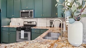 Una cocina con gabinetes verde mar, electrodomésticos de acero inoxidable, encimeras de granito y un jarrón blanco con hojas verdes.