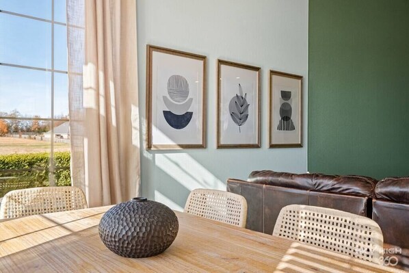 A dining area with a wooden table, wicker chairs, a brown leather couch, and three framed artworks on the wall.