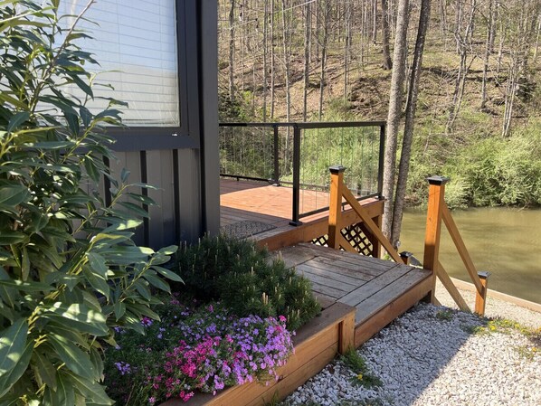 A wooden deck with a railing, surrounded by greenery and flowers. - The Hideaway on Tacketts Shipping Container (Hurricane)
