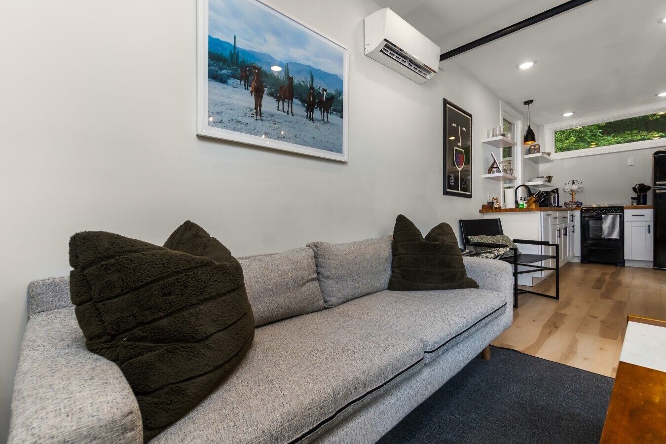 A modern living room with a grey sofa, dark cushions, a flat-screen TV, and a kitchen in the background.