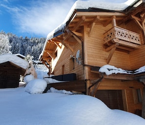 A wooden chalet with a balcony, surrounded by snow-covered trees and a clear sky.