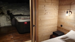 Een houten interieur met een bed, een klein krukje en een zichtbare hottub door een geopende deur.