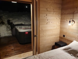 A wooden interior with a bed, a small stool, and a hot tub visible through an open door.
