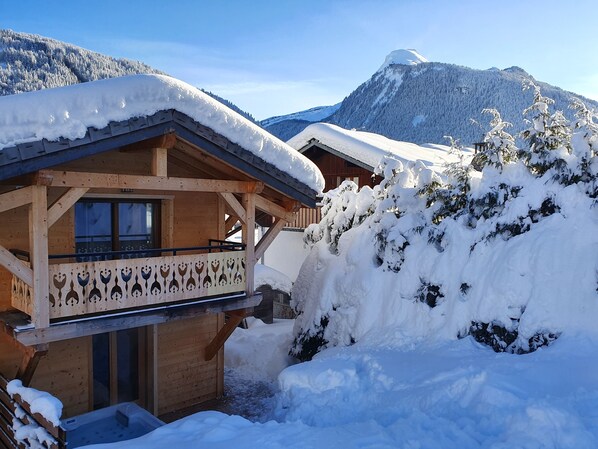 A wooden chalet with a balcony, surrounded by snow-covered trees and mountains.