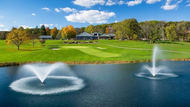 Un campo de golf con club house, un estanque con fuentes y árboles de colores otoñales.