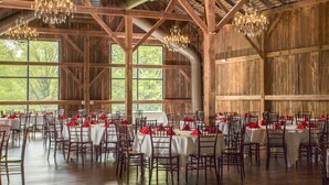Un amplio salón con vigas de madera y candelabros, mesas dispuestas con manteles blancos y servilletas rojas, y grandes ventanales con vistas a árboles.