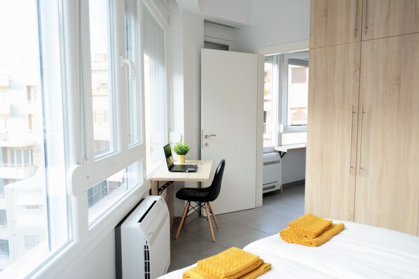 Un dormitorio moderno con una cama, un escritorio y una silla. Hay una ventana con vistas a edificios.