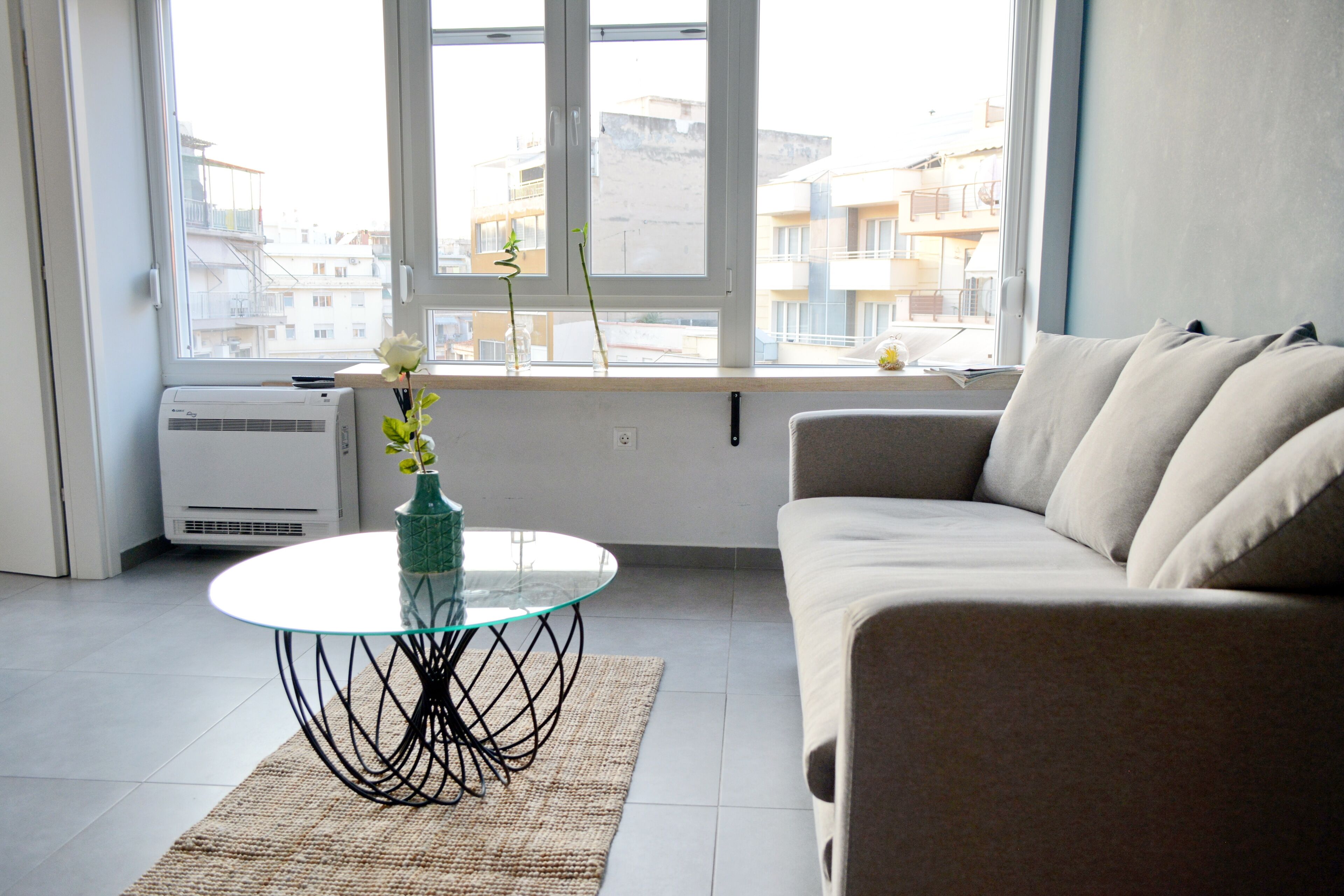 A living room with a glass table, a beige sofa, and a view of buildings through a large window.