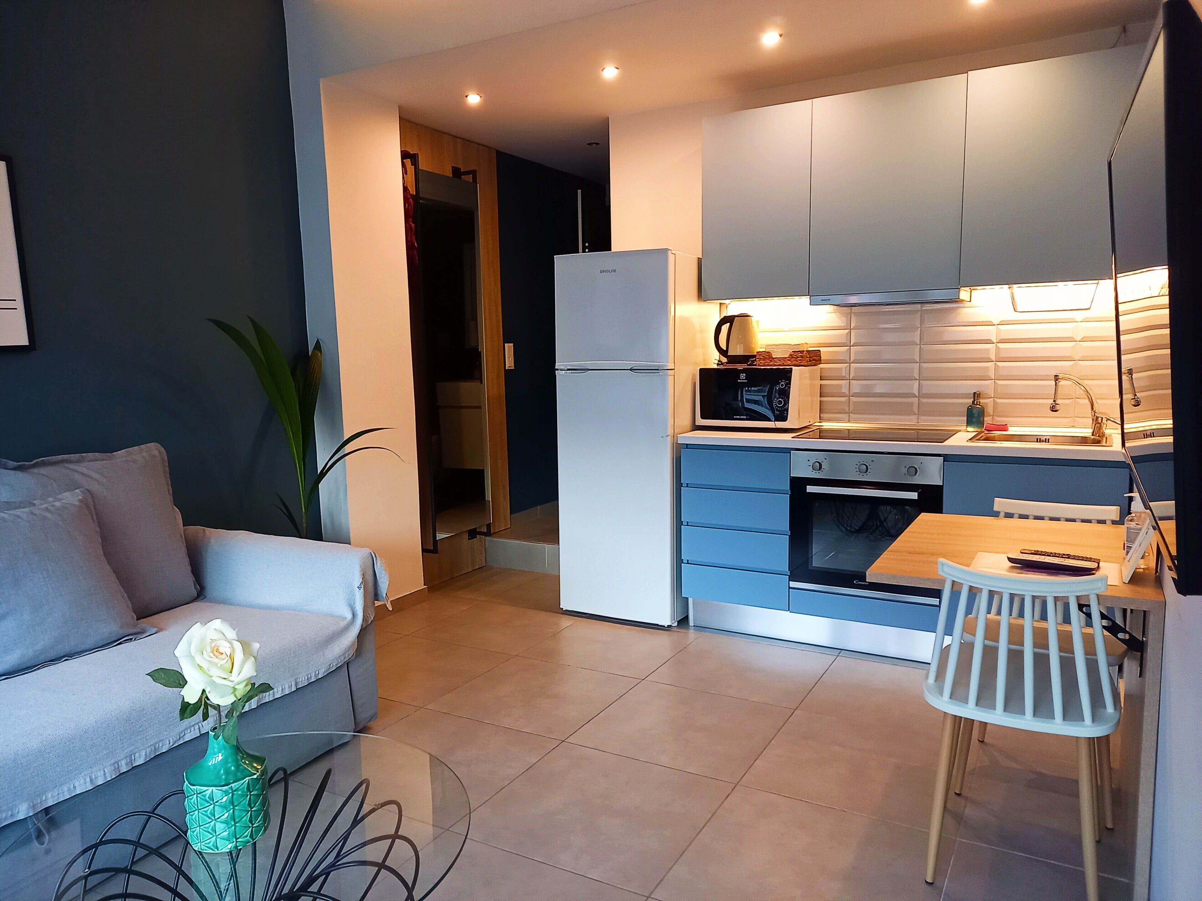 Een kleine keuken met een koelkast, magnetron en fornuis, een eettafel met stoelen, en een bank met een bloevas.