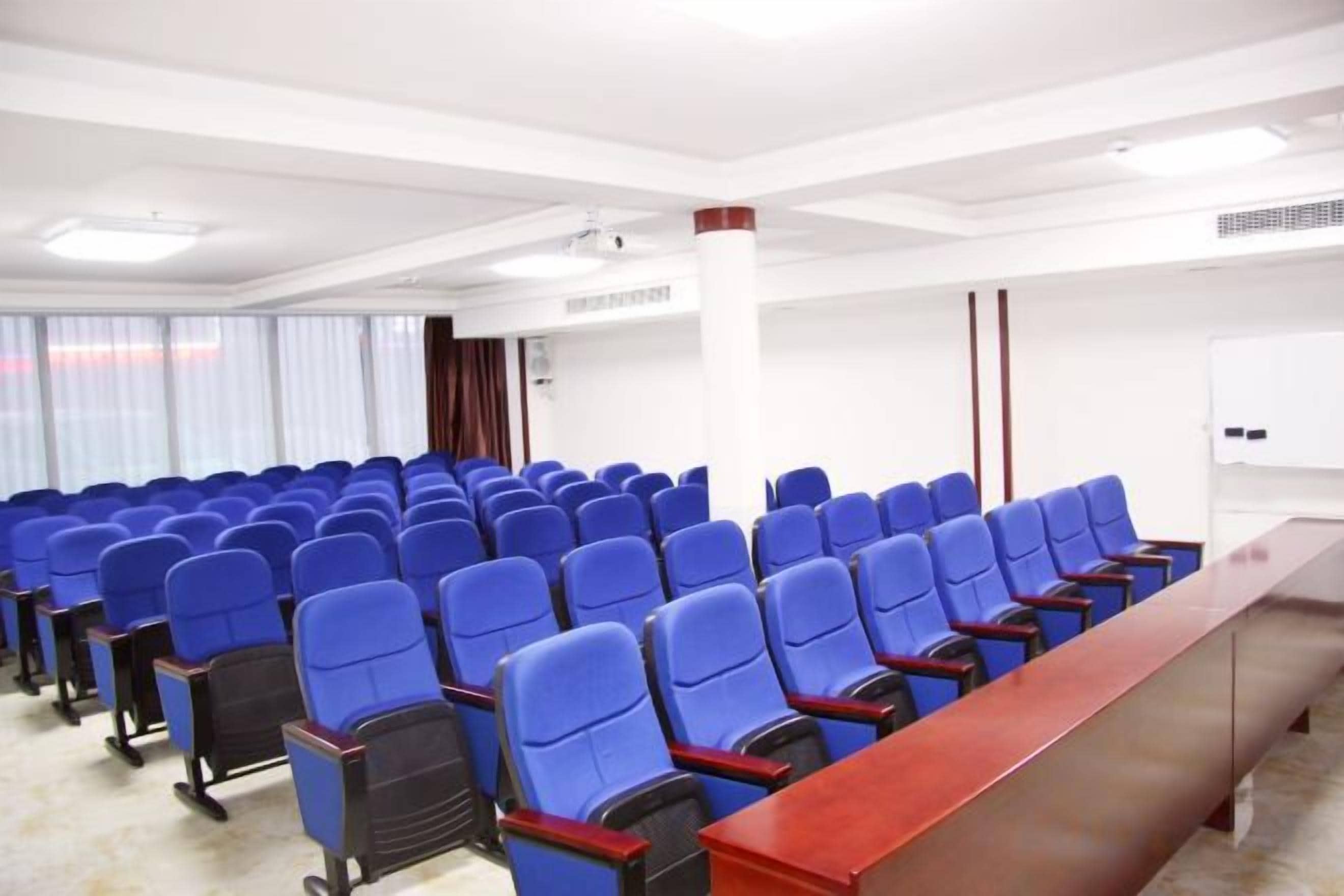 Sala de conferência com fileiras de cadeiras azuis, uma mesa de madeira comprida e um quadro branco.
