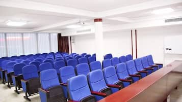 Sala de conferência com fileiras de cadeiras azuis, uma mesa de madeira comprida e um quadro branco.