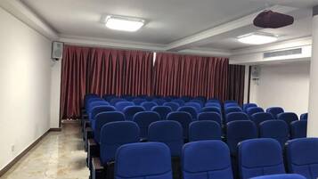 A conference room with rows of blue chairs, a red curtain backdrop, and ceiling lights.