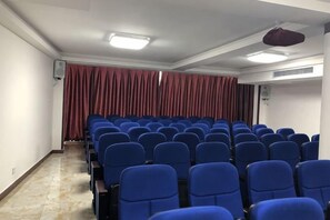 Une salle de conférence avec des rangées de chaises bleues, un rideau rouge en arrière-plan et des luminaires au plafond.