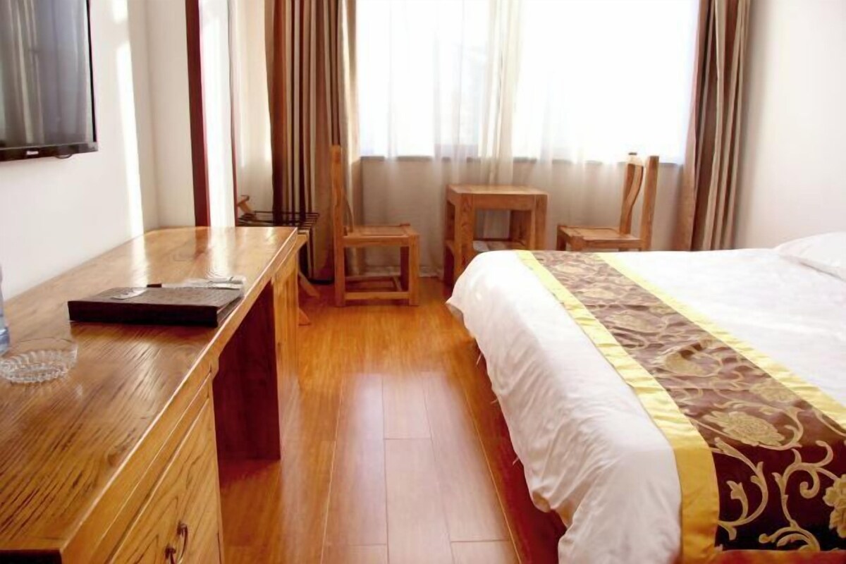 Hotelkamer met een houten bureau, twee stoelen, een bed met een beddegoed met dessin en een raam met gordijnen.